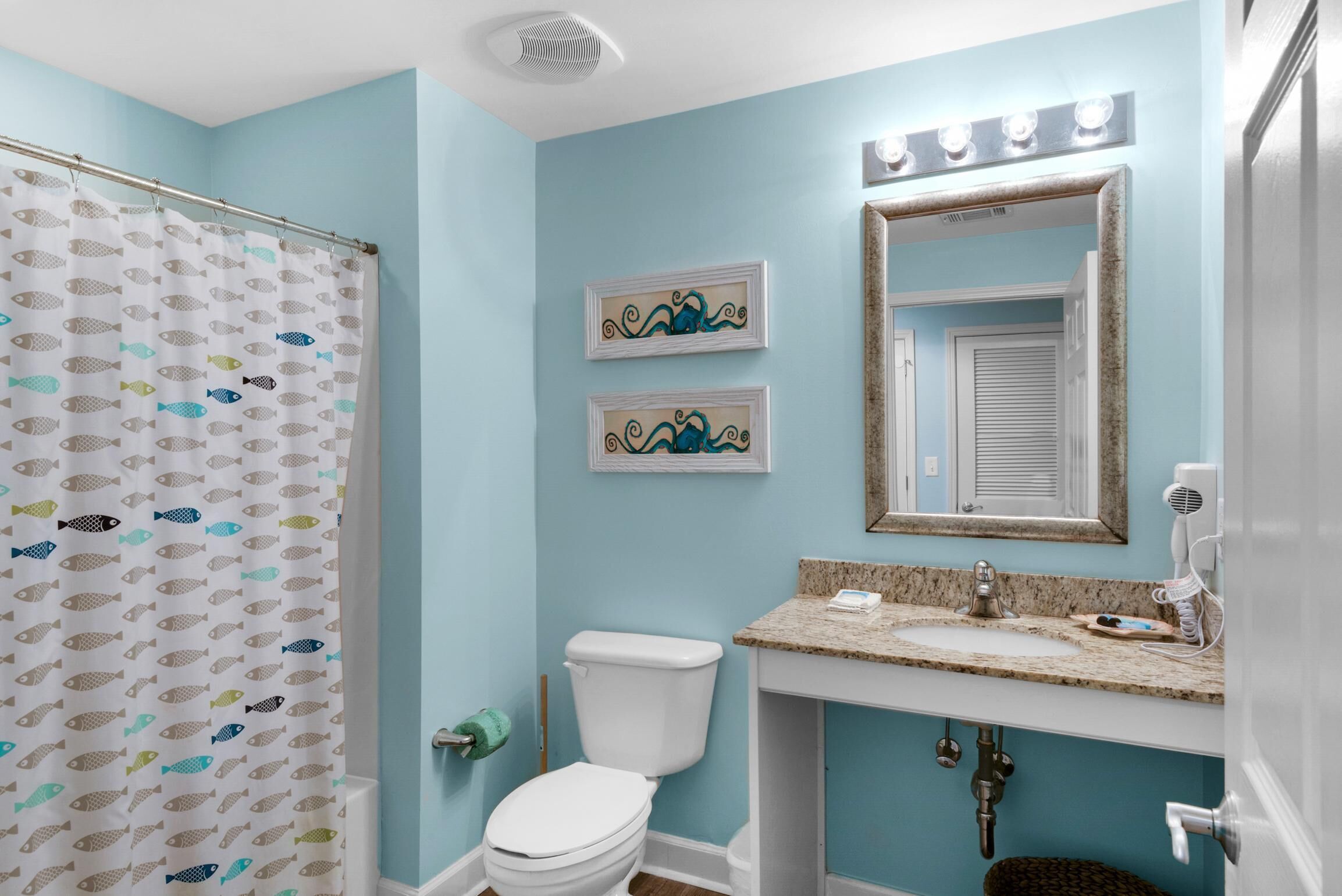 A bathroom with a light blue wall, a white toilet, a shower curtain with fish patterns, a sink with a granite countertop, and two framed wall art pieces.