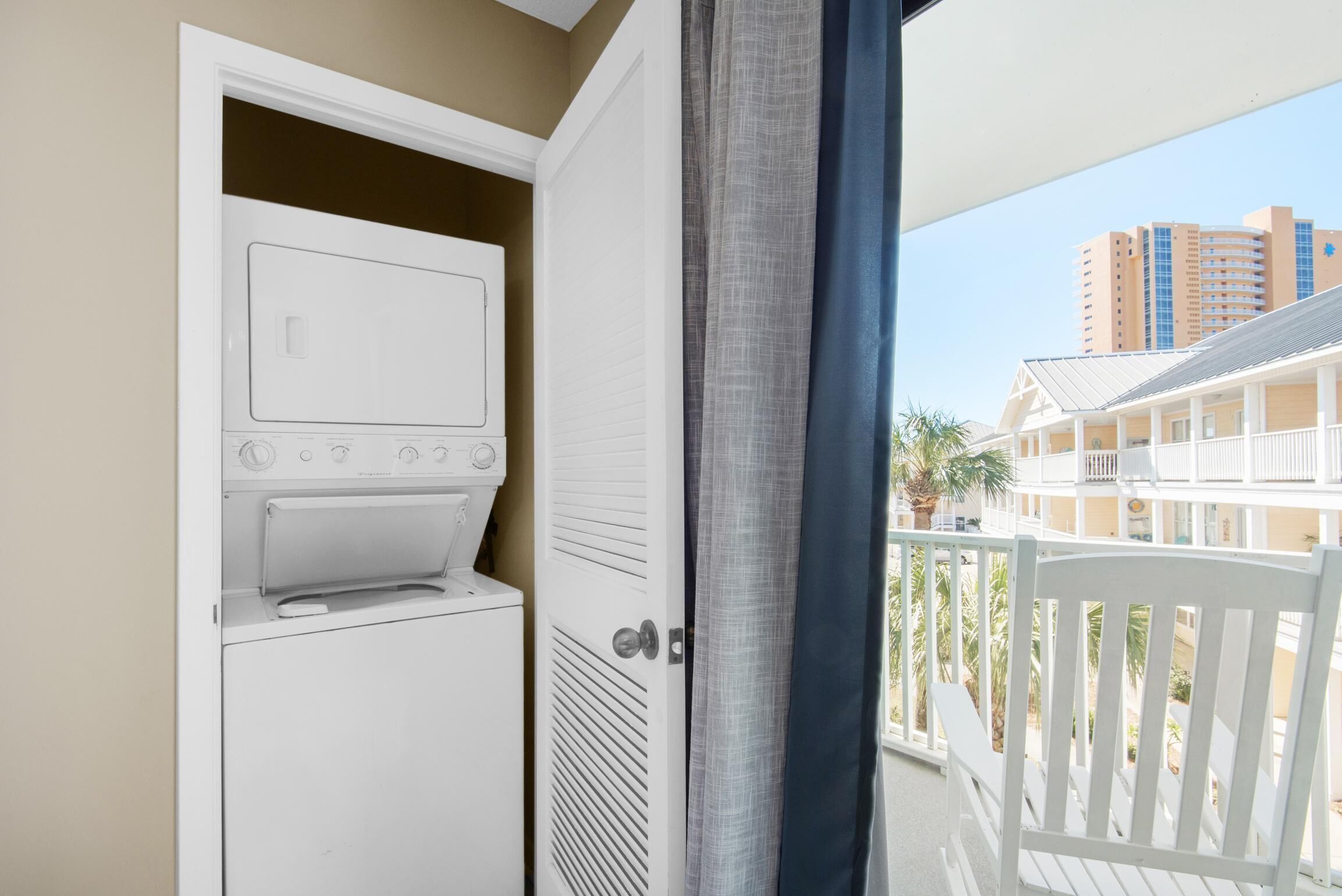 A laundry room with a washer and dryer, a door leading to a balcony with a view of a building and palm trees.