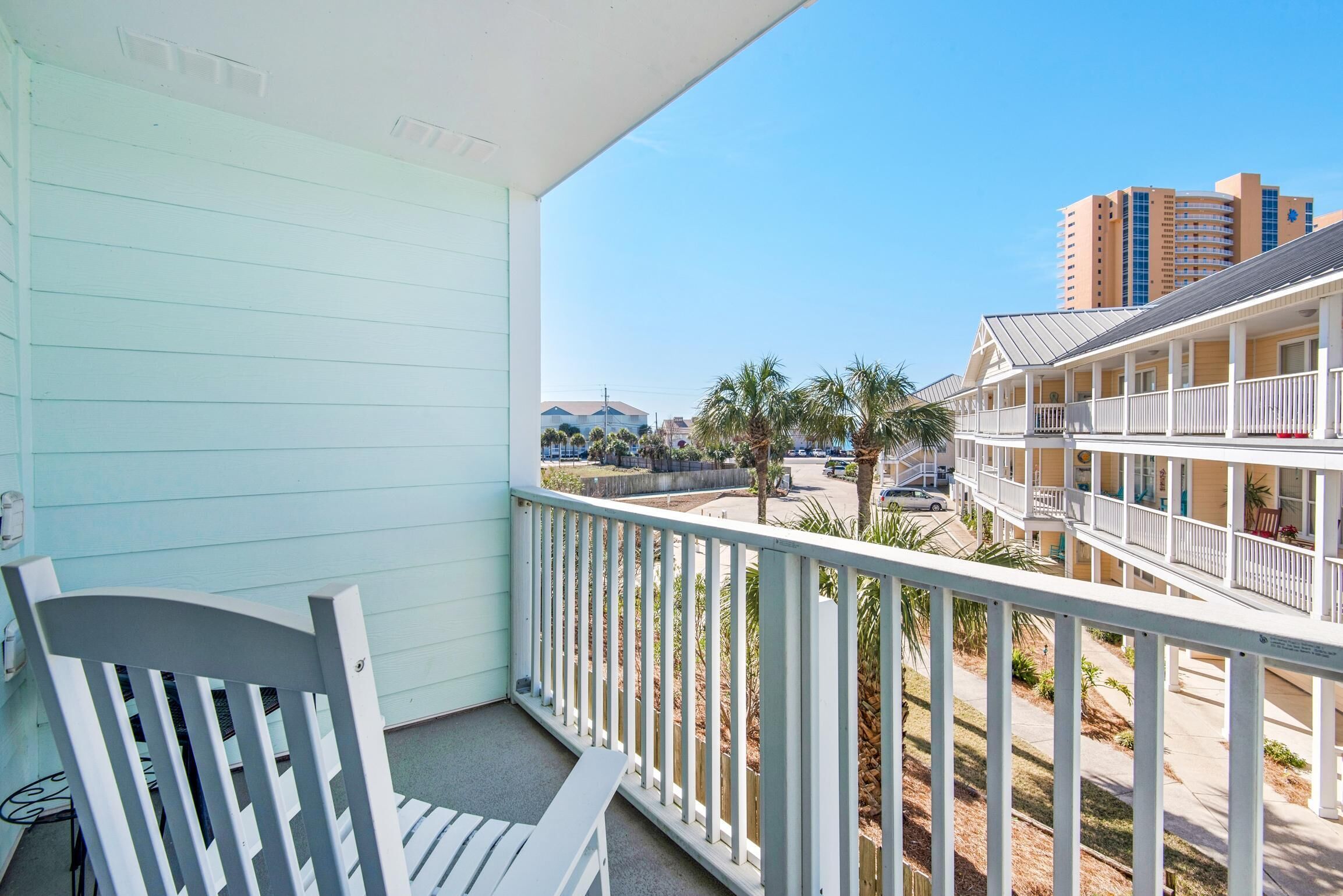 A balcony with a white chair, overlooking a street with buildings and palm trees.