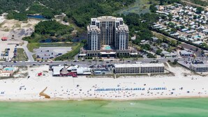 An aerial view of a coastal hotel with a central building, a beach with umbrellas, and a parking lot. - Origin at Seahaven 825 (Panama City Beach)