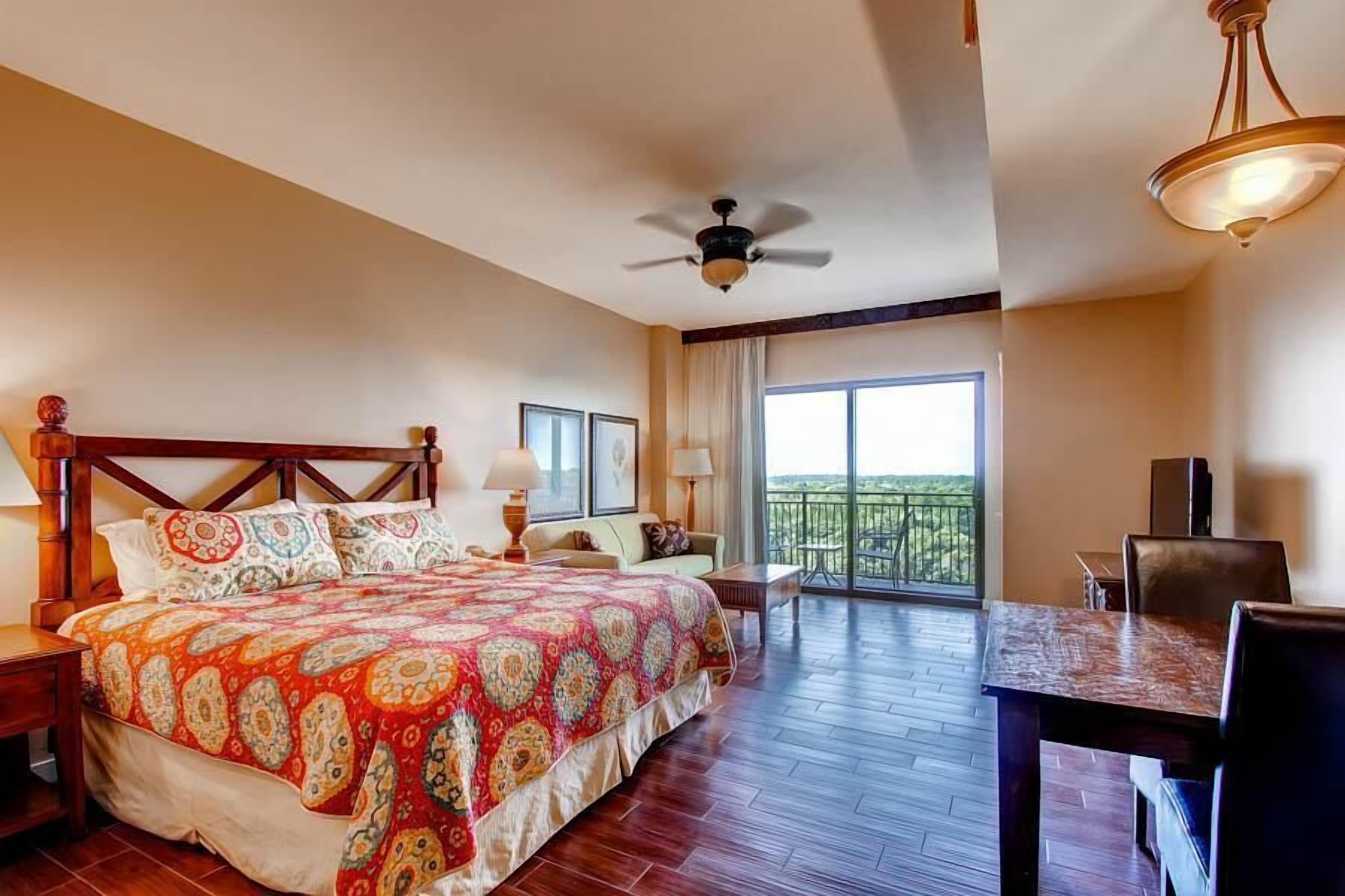 Una habitación de hotel espaciosa con una cama grande, un balcón con vistas, una mesa de comedor y un ventilador de techo.