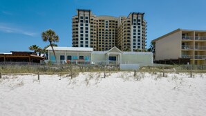 A beachfront hotel with a white building, a sandy beach in the foreground, and a tall hotel building in the background. - Origin at Seahaven 825 (Panama City Beach)