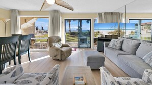 A spacious living room with a sofa, armchair, and coffee table. A ceiling fan and a TV are visible. The room has large windows with a view of the beach.