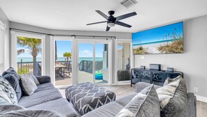 Ein Wohnzimmer mit grauem Sofa, gemusterten Kissen, Deckenventilator und großem Fenster mit Blick auf einen Strand.