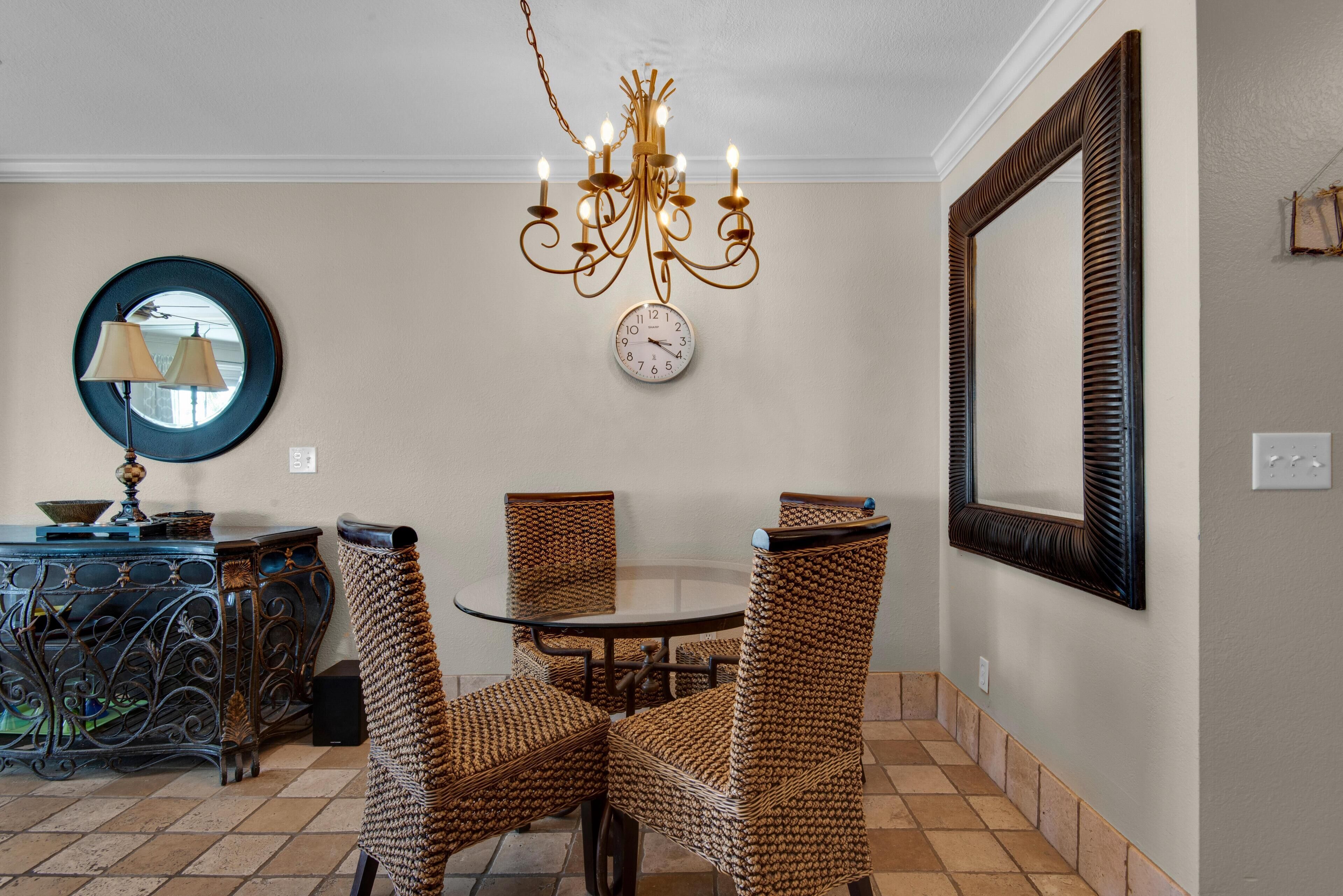 A dining area with wicker chairs, a round table, a chandelier, a wall clock, and a mirror.