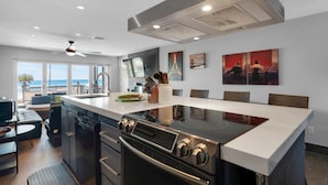 Een moderne keuken met een groot eiland, een ingebouwde oven en roestvrijstalen apparatuur. Er staan eetstoelen en een tafel met uitzicht op de oceaan.