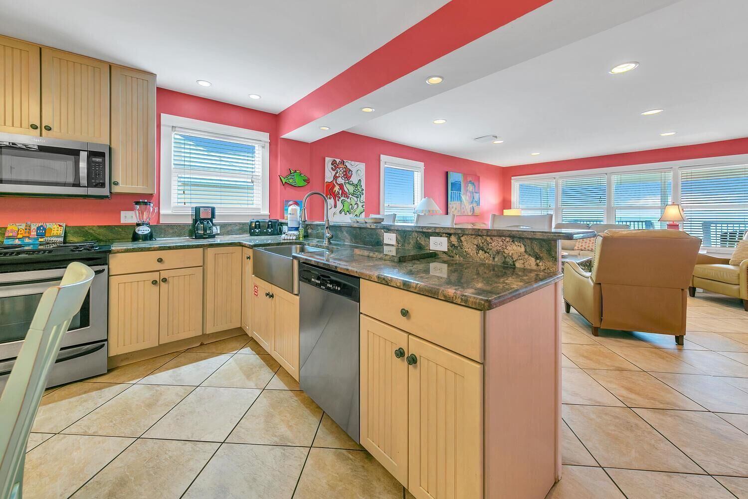 A modern kitchen with wooden cabinets, a built-in microwave, and a dishwasher. There are two windows with blinds, a red accent wall, and a dining area with a brown leather chair.
