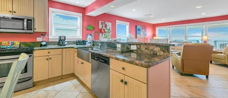 A modern kitchen with wooden cabinets, a built-in microwave, and a dishwasher. There are two windows with blinds, a red accent wall, and a dining area with a brown leather chair.