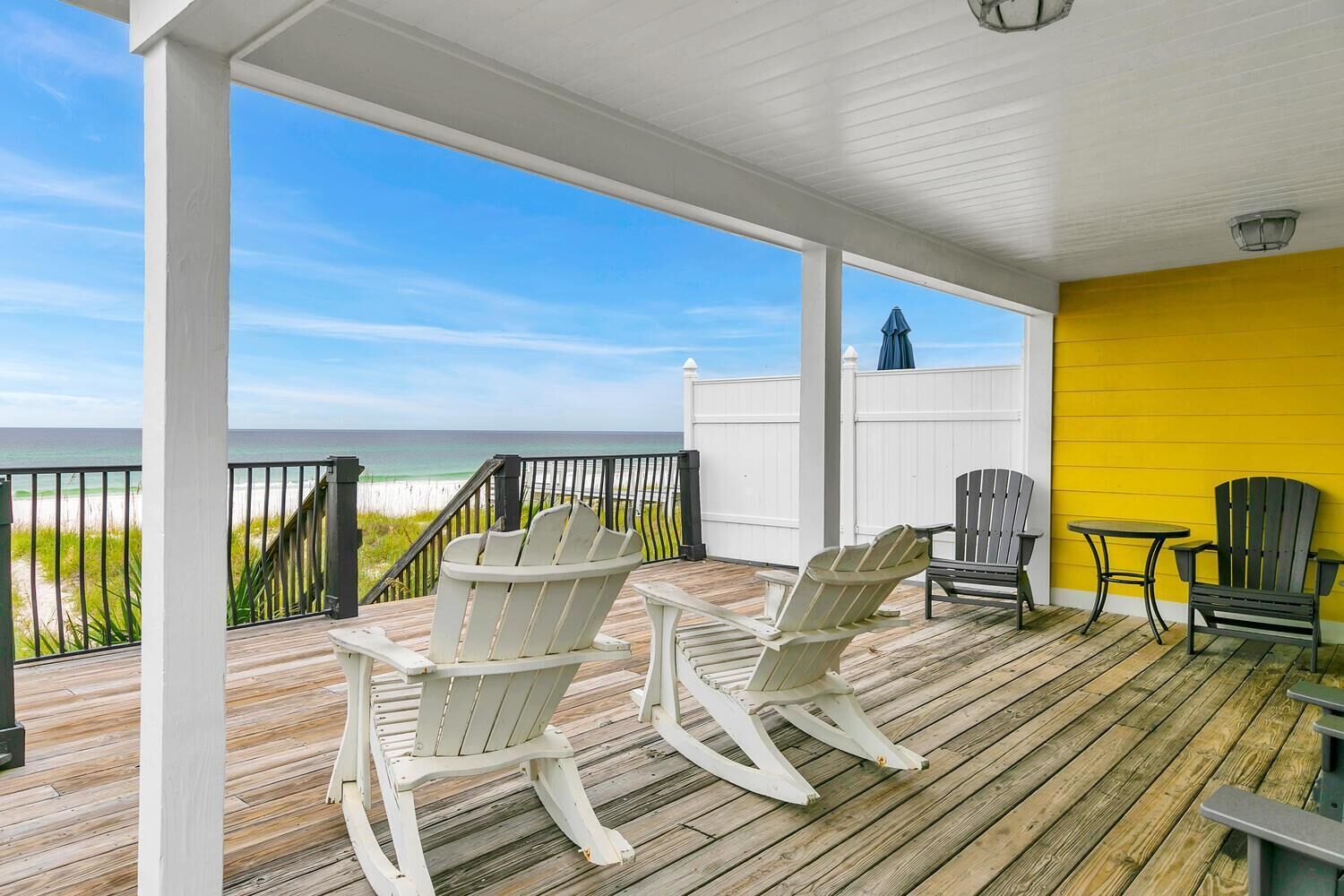 A wooden deck with two rocking chairs and a table, overlooking a beach with a white fence and a yellow wall.
