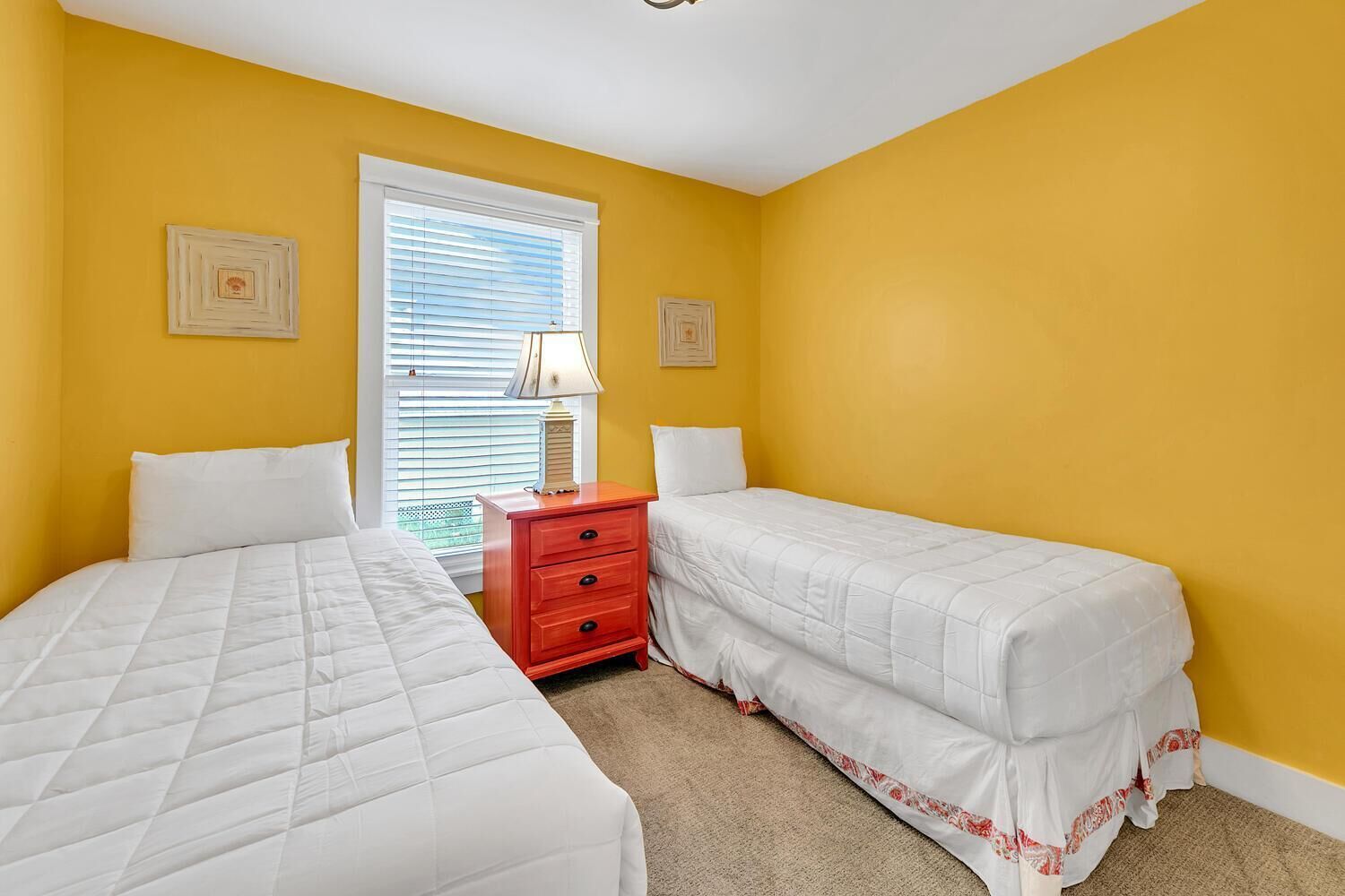 A bedroom with two beds, a red nightstand, and a window with blinds.