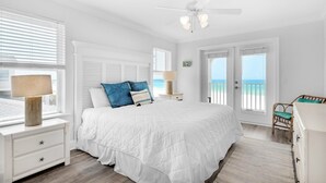 Ein Schlafzimmer mit einem großen Bett, einem Deckenventilator, einem Nachttisch mit Lampe, einer Kommode und Meerblick durch eine Tür.