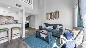 A modern living room with a grey sofa, a coffee table, and a blue rug. There are two wooden stools and a kitchen area with white cabinets. - Endless Summer F9 (Panama City Beach)