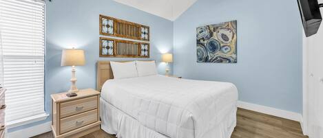 Un dormitorio con una cama, dos mesitas de noche con lámparas, un ventilador de techo y una obra de arte colgada en la pared.