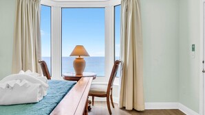 Una habitación de hotel con un amplio ventanal, una cama con edredón blanco, una mesita de noche de madera, una lámpara y una silla.