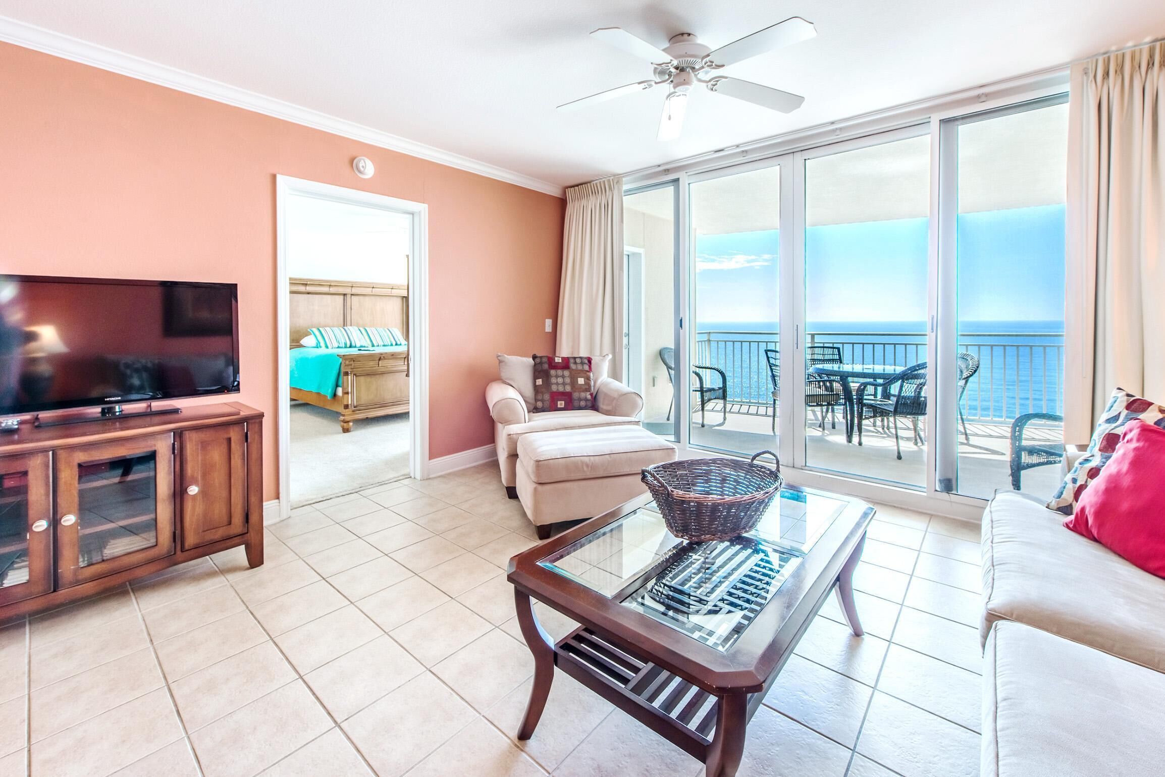 A living room with a television, a glass-top coffee table, a sofa, and a balcony with a view of the ocean.