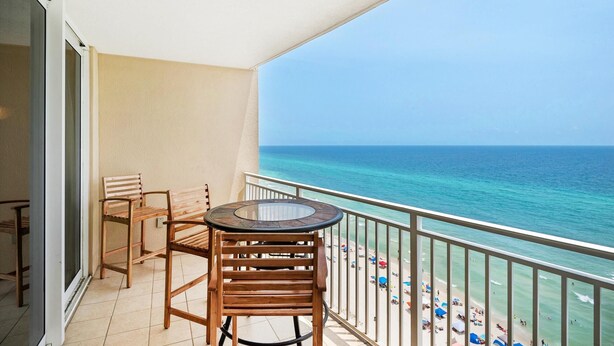 A balcony with a wooden table and chairs, overlooking a beach with umbrellas and people. - Emerald Beach 1627 (Panama City Beach)