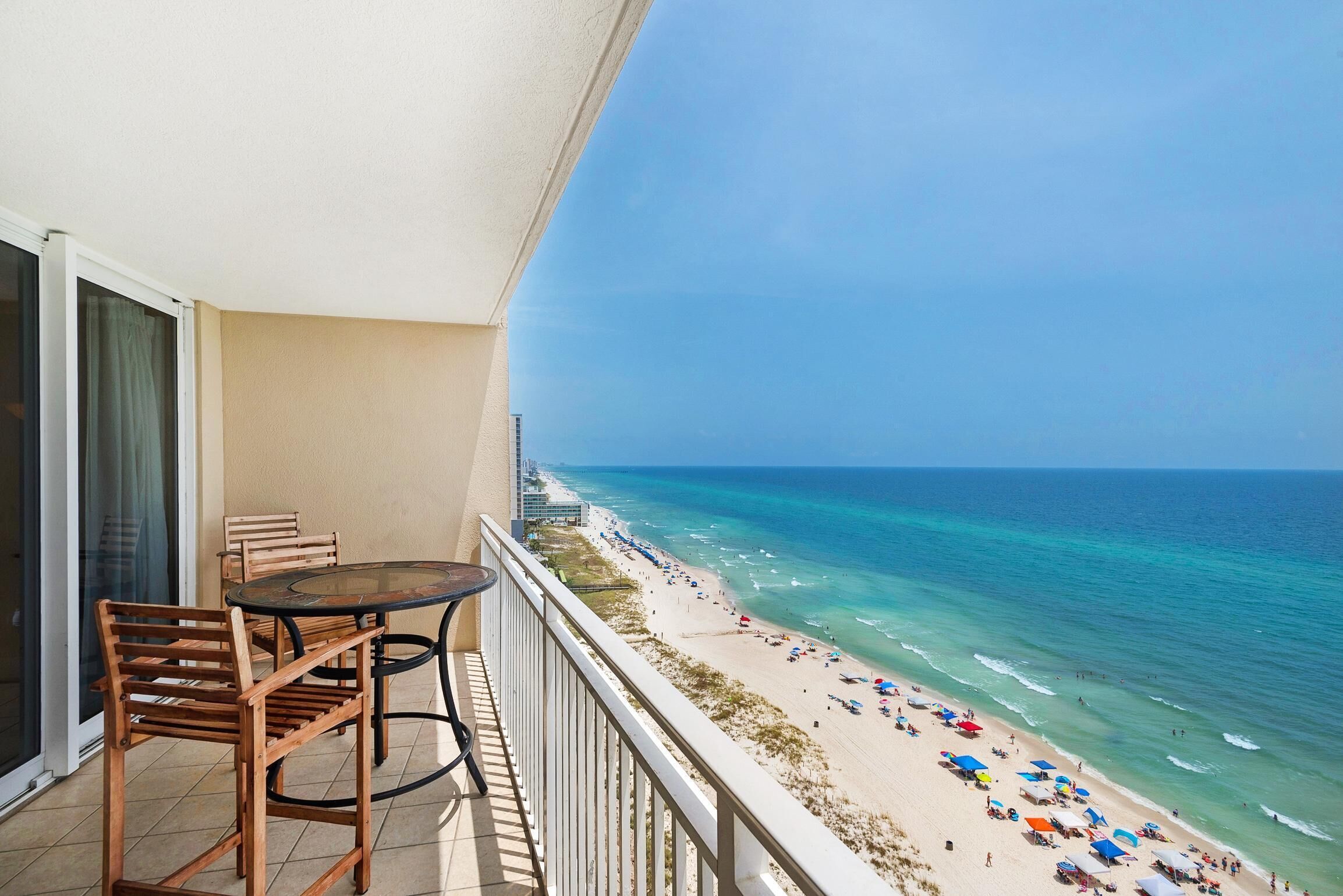 A balcony with a table and chairs overlooking a beach with umbrellas and people.