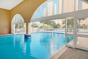 An indoor swimming pool with clear glass walls, a tiled floor, and a view of palm trees and buildings outside. - Celadon 703 (Panama City Beach)