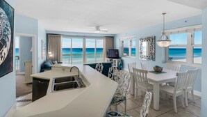 Une cuisine avec vue sur l’océan, un coin repas avec des chaises blanches et un téléviseur posé sur un meuble.