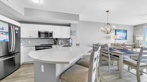 A modern kitchen with a central island, stainless steel appliances, and a dining area with wooden chairs and a chandelier. - Celadon 1902 "aloha No Ka Oi" (Panama City Beach)