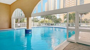 An indoor swimming pool with clear blue water, large arched windows, and a view of palm trees and buildings outside. - Celadon 1902 "aloha No Ka Oi" (Panama City Beach)