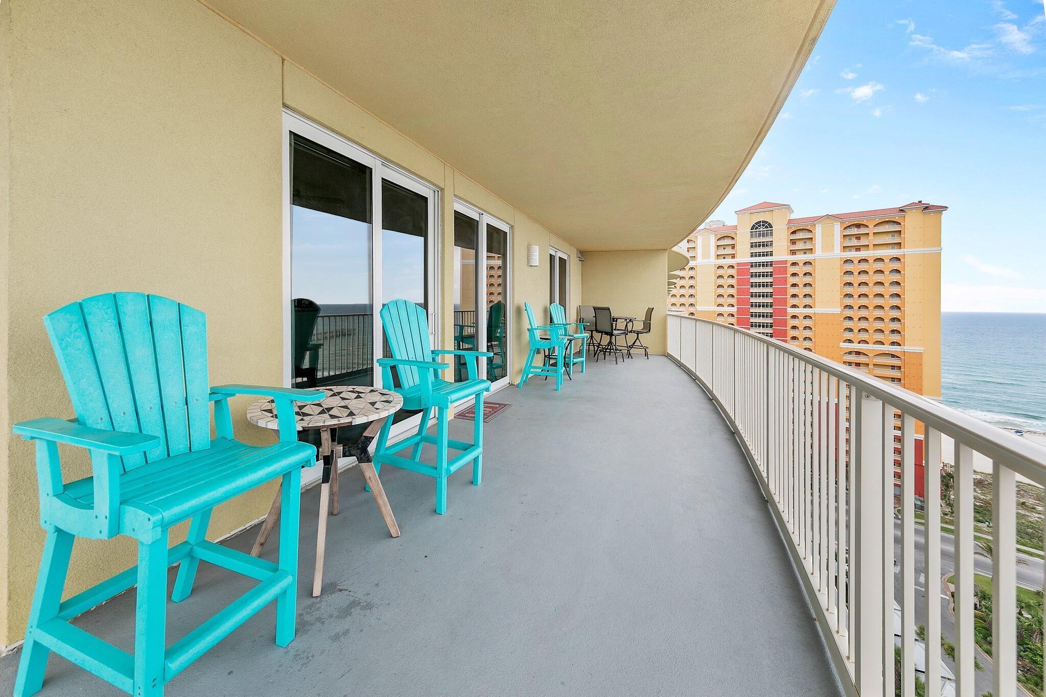 A balcony with turquoise chairs and a small table, overlooking a body of water and a hotel building.