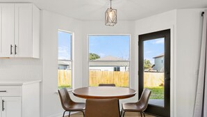 A kitchen with white cabinets, a round dining table with four chairs, and a view of a backyard through large windows. - Casa Blanca  (Kyle)