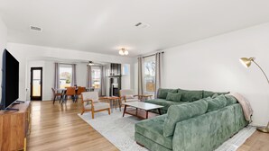 A modern living room with a green sofa, wooden coffee table, and a TV on a wooden stand. There is a dining area with chairs and a table, and a kitchen area with cabinets and a stove. - Casa Blanca  (Kyle)