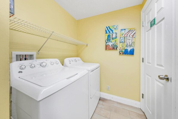 A laundry room with two white front-loading washers and dryers, a metal drying rack, and two colorful paintings on the wall.