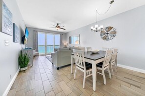 A modern living room with a dining area, a ceiling fan, and a view of the ocean through large windows. - Calypso 2-1102 West (Panama City Beach)