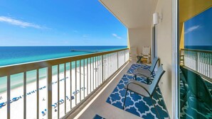 A balcony with a view of the beach and ocean, featuring a railing, a chair, and a patterned rug.