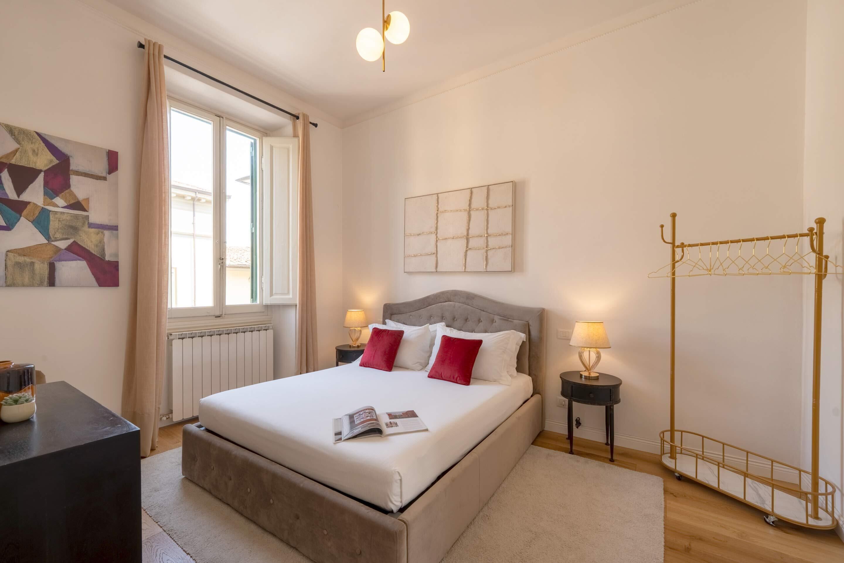 A bedroom with a bed, a window, a radiator, a bedside table with a lamp, and a wall art piece.