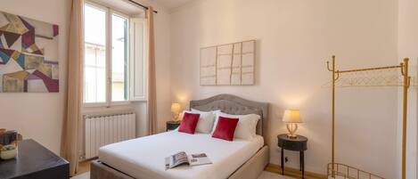 A bedroom with a bed, a window, a radiator, a bedside table with a lamp, and a wall art piece.