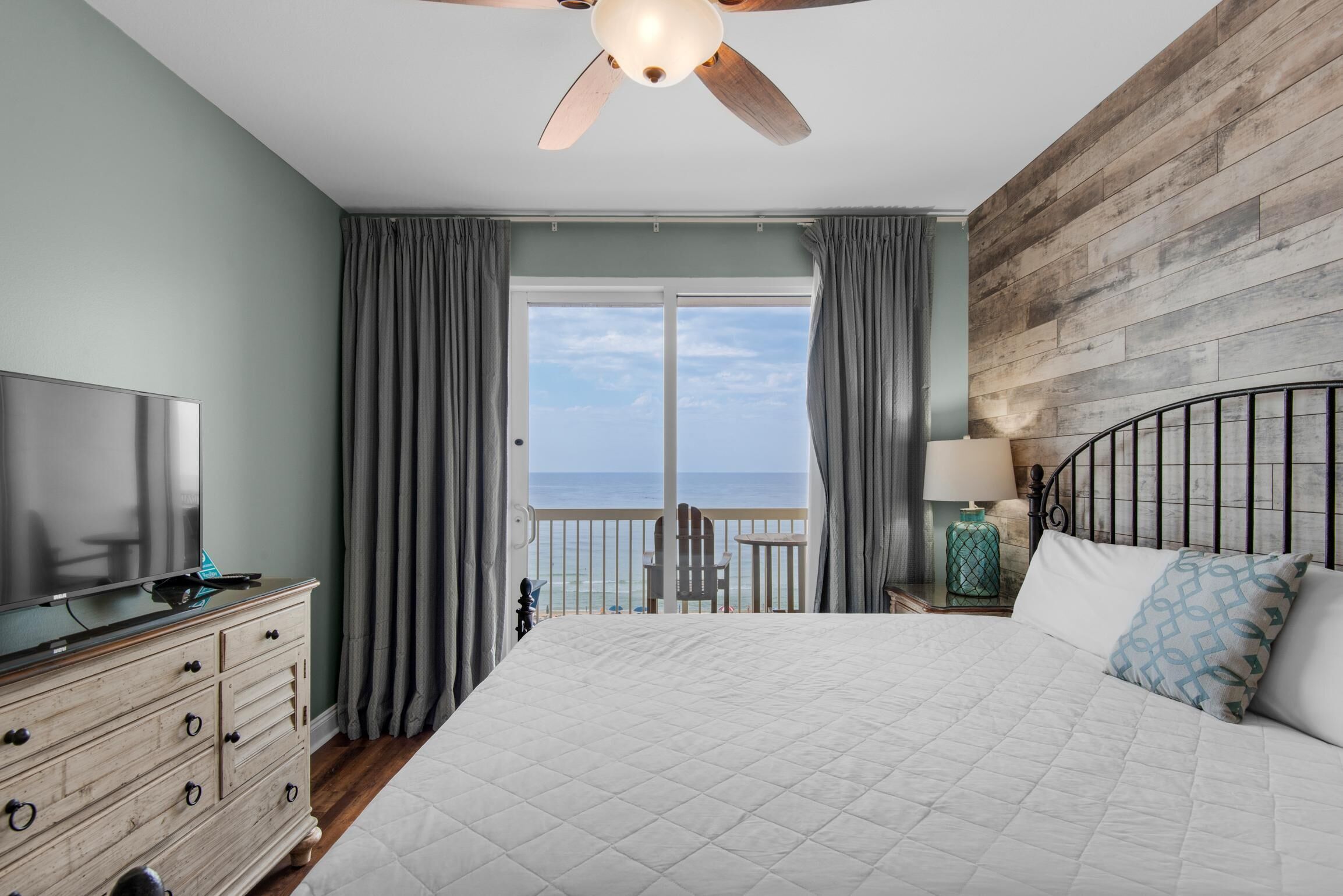 Une chambre avec un grand lit, un téléviseur posé sur une commode, un ventilateur de plafond et une vue sur l’océan par la fenêtre.
