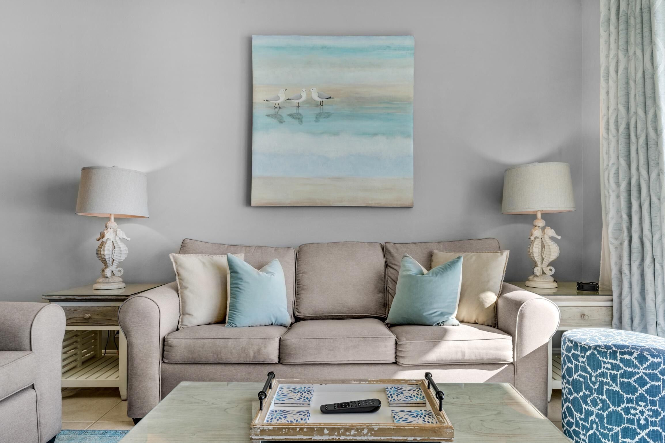 A living room with a beige sofa, two side tables with lamps, a coffee table with a tray, and a painting of seagulls on the wall.