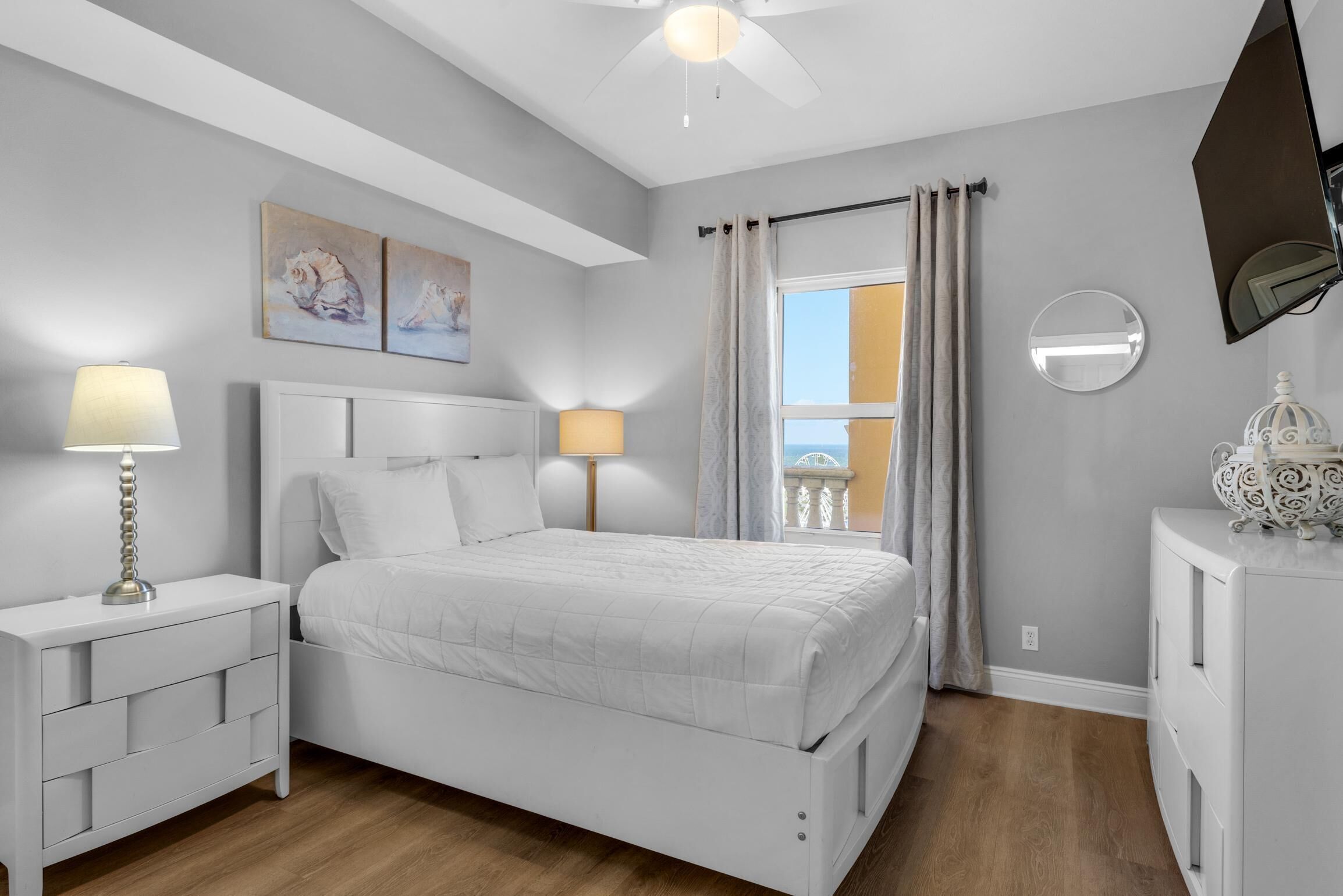 Un dormitorio con cama, mesitas de noche, ventilador de techo, ventana con cortinas y una obra de arte en la pared.