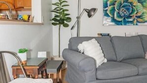 Un espacio habitable compacto con kitchenette, un sofá gris, una mesita, una silla, una lámpara de pie y un cuadro de una flor.