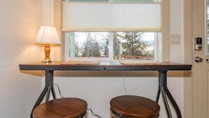 Une pièce avec une table et deux tabourets, une lampe et une fenêtre avec des stores.