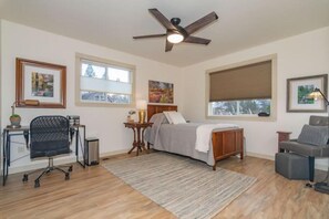 A bedroom with a bed, a desk, a chair, a ceiling fan, and large windows.