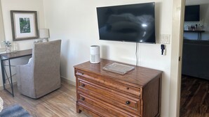 Ett rum med en platt-TV som är monterad på väggen, en byrå i trä med lådor, en stol, ett skrivbord med en lampa och en inramad tavla på väggen.