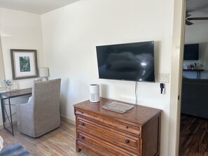 Une pièce dotée d’un téléviseur à écran plat fixé au mur, d’une commode en bois avec des tiroirs, d’une chaise, d’un bureau avec une lampe et d’un tableau encadré accroché au mur.