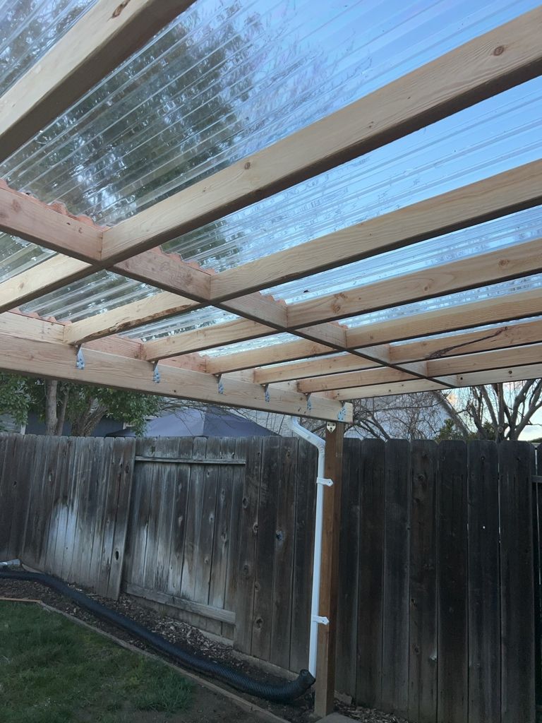 Una estructura de pérgola de madera con techo transparente, rodeada por una valla de madera.