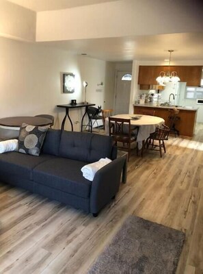 A modern living room with a dark grey sofa, a dining table with chairs, and a kitchen area with wooden cabinets and a white refrigerator. - Townhome 2 bed, 2 bath, fenced backyard & garage (Central Point)
