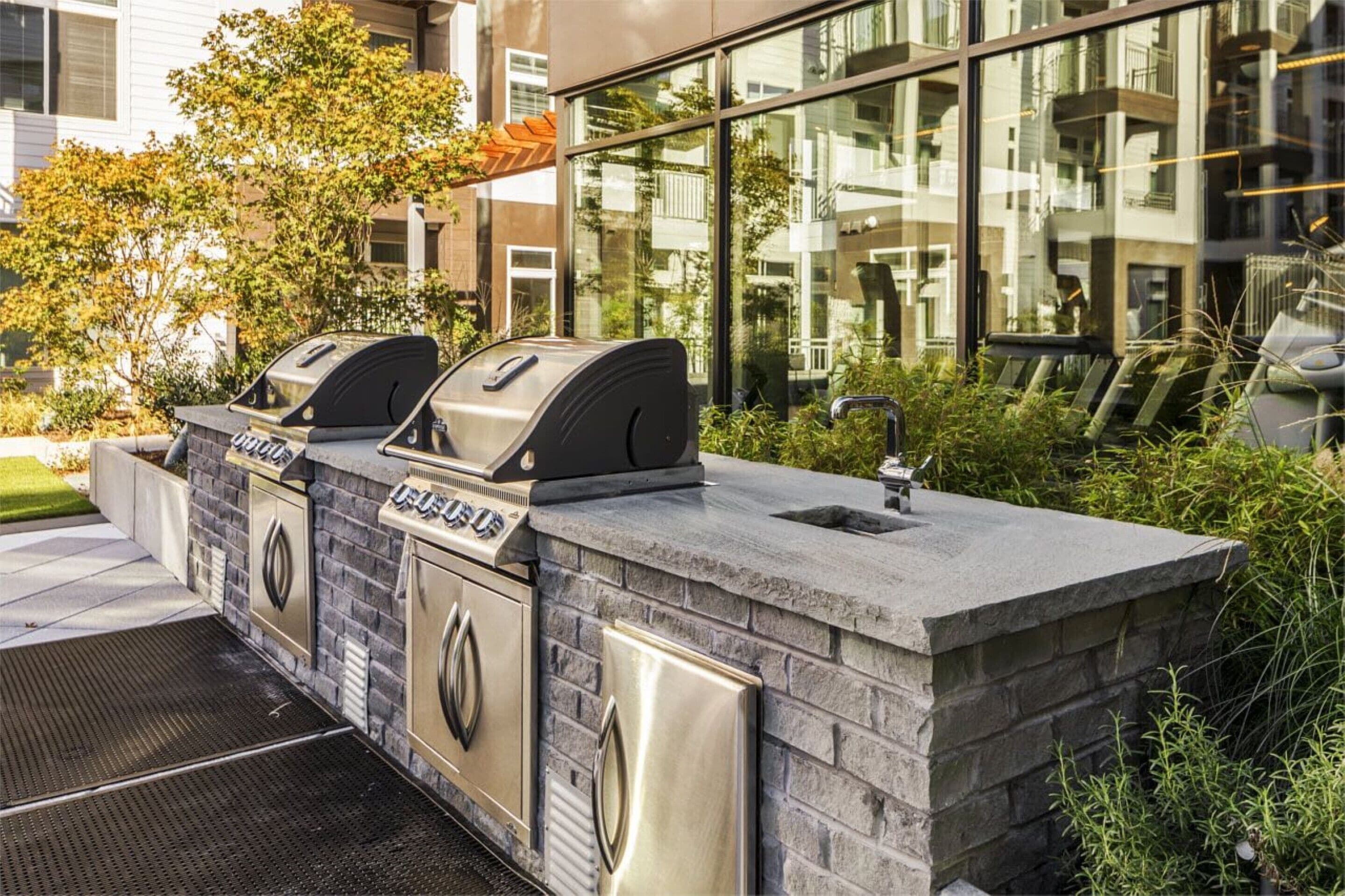 An outdoor kitchen with two grills, a sink, and a stone countertop, surrounded by greenery and a modern building in the background.