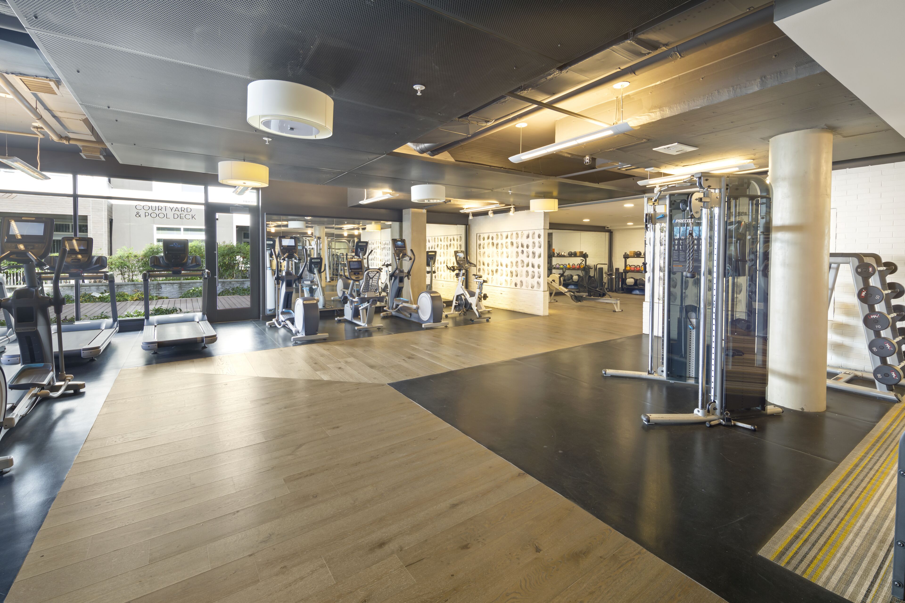 A gym with various exercise equipment, a wooden floor, and large windows.
