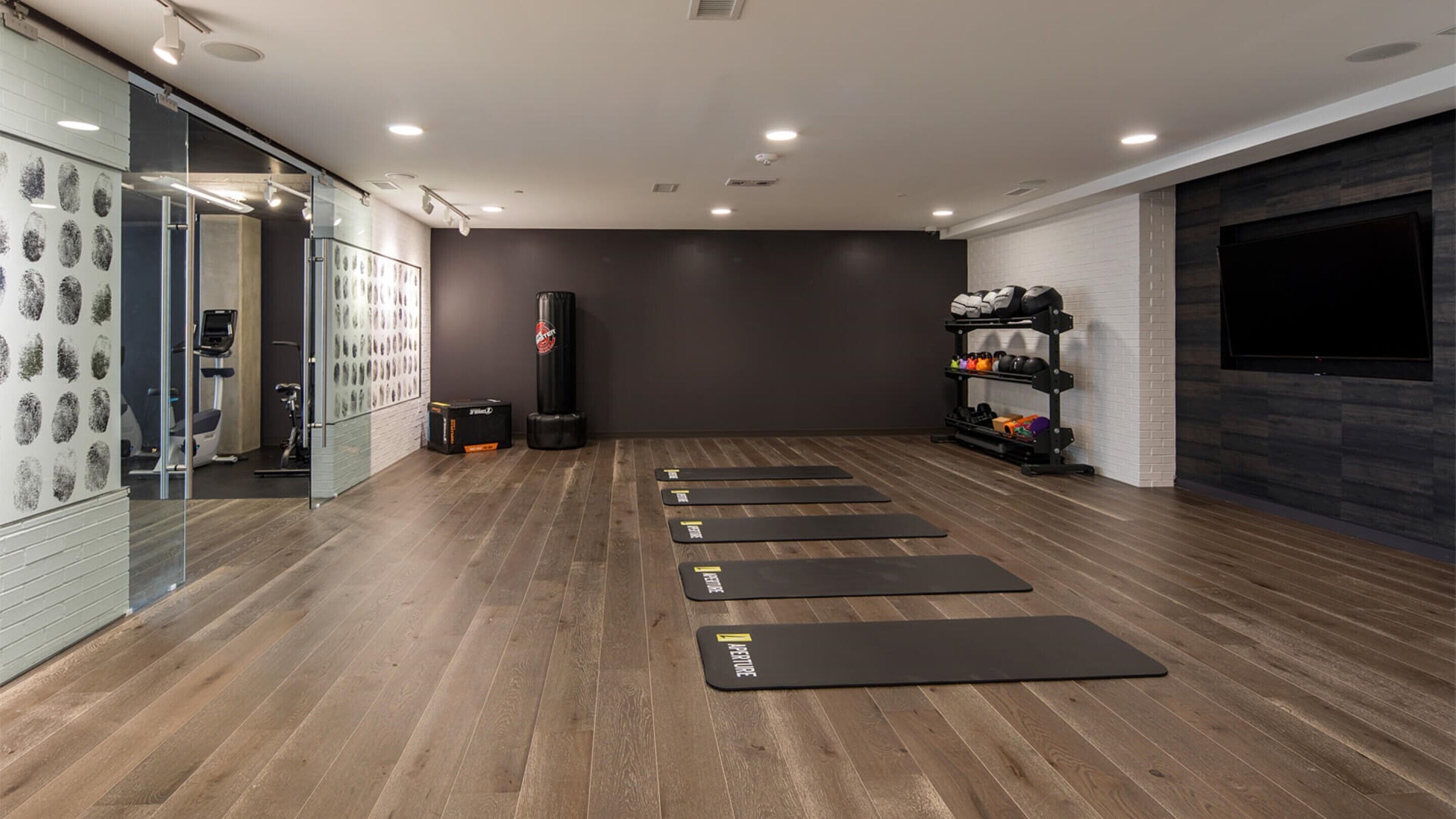 Un gimnasio moderno con suelo de madera, colchonetas de ejercicio, un saco de boxeo y un televisor montado en la pared.