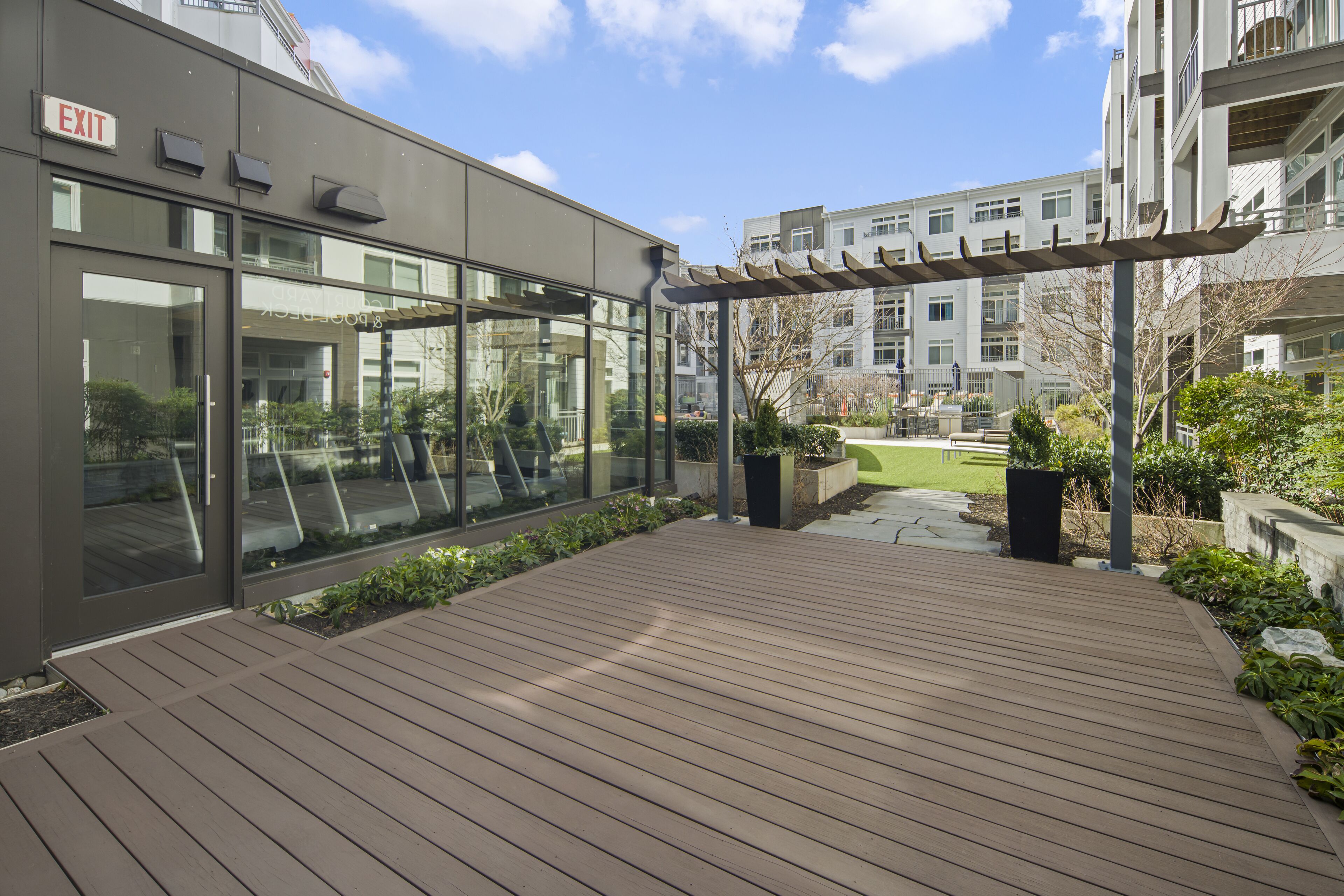 Una terraza moderna al aire libre con una pérgola, una entrada de vidrio y vistas de edificios residenciales al fondo.