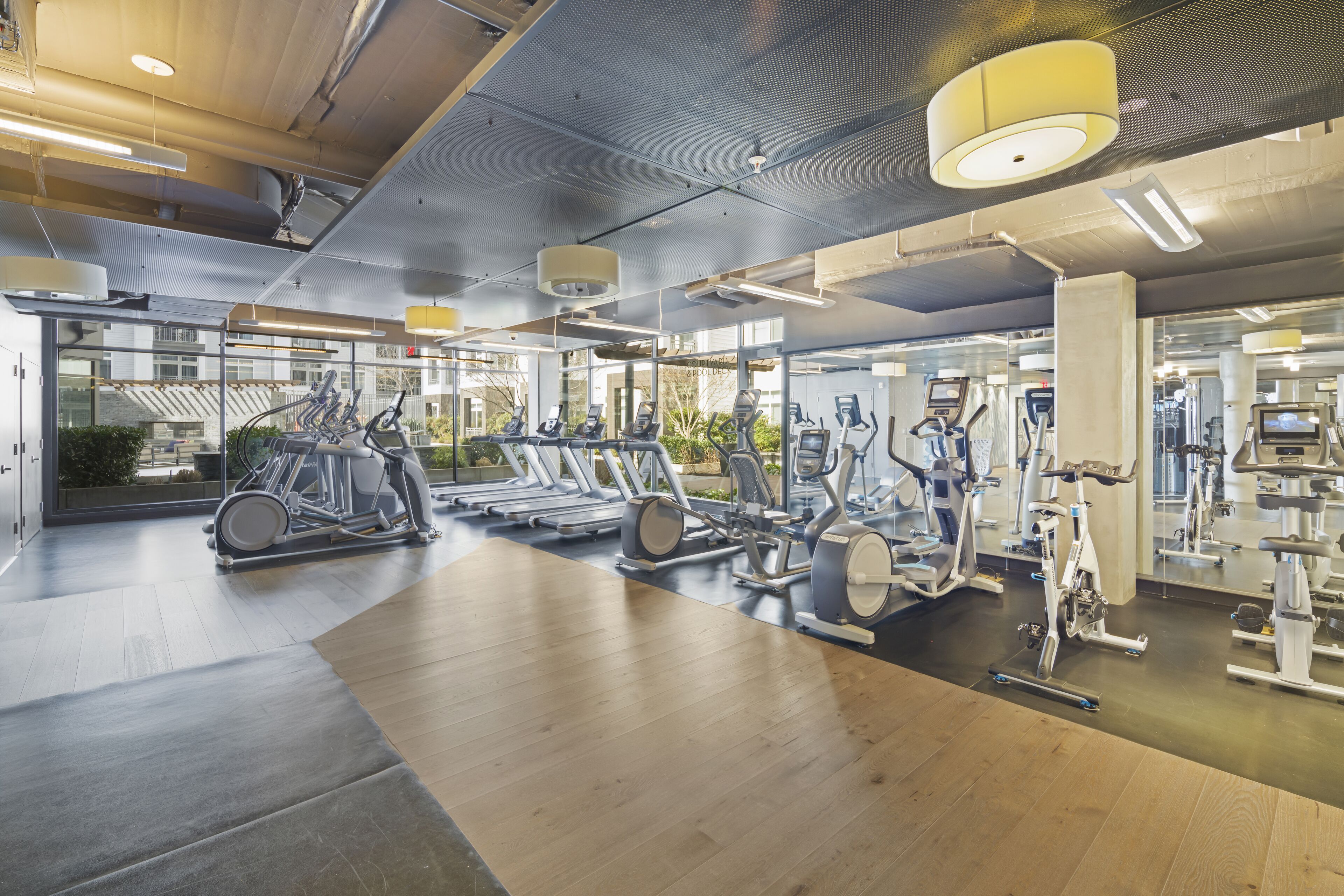 Academia moderna com diversos equipamentos de exercício, grandes janelas e piso de madeira.