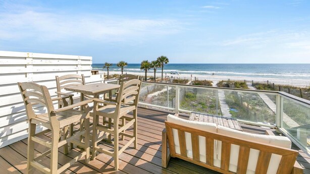 A wooden deck with a table and chairs, overlooking a beach with palm trees and a clear sky. - Beachside West 13 (Panama City Beach)