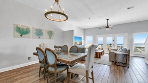 A spacious dining area with a wooden table and chairs, a chandelier, and a view of the beach through large windows. - Beachside West 13 (Panama City Beach)